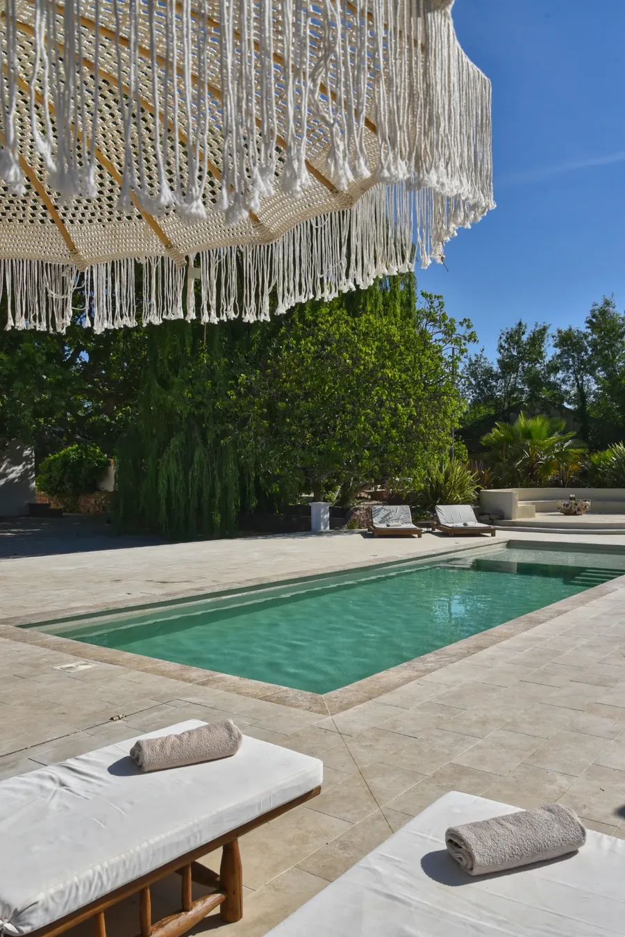 La même piscine vue sous un parasol en macramé, eau limpide et végétation